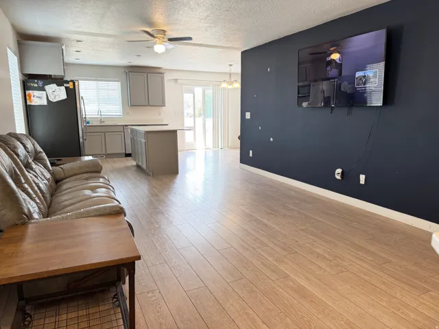 Open-concept living space featuring wood-finish flooring, a contemporary kitchen with light gray cabinetry and an island, a dark accent wall, and a ceiling fan with integrated lighting