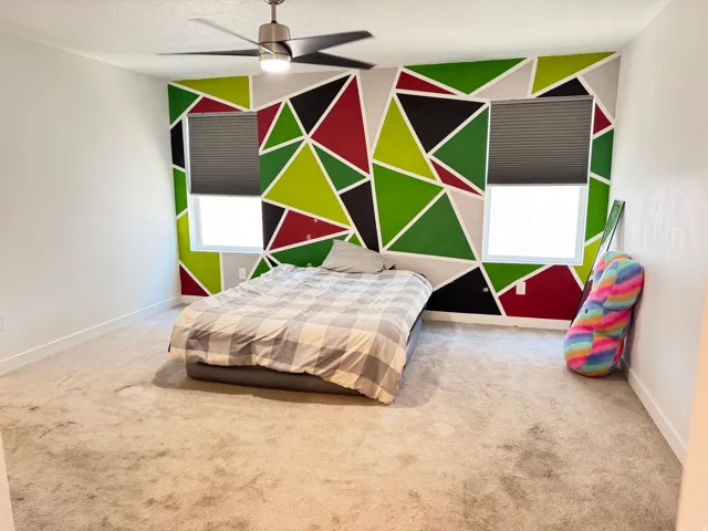 Bedroom featuring a geometric accent wall with dual windows, neutral carpeting, and a ceiling fan with integrated lighting