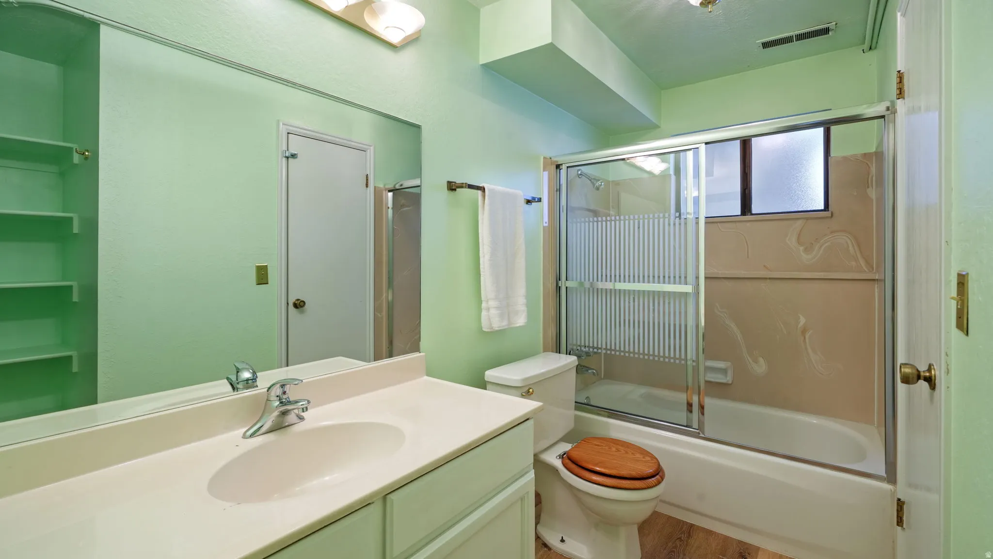 Bathroom with a light green color palette featuring a vanity with an integrated sink and chrome faucet, a large wall-mounted mirror, built-in shelving, and a tub/shower combination with a sliding glass enclosure and window