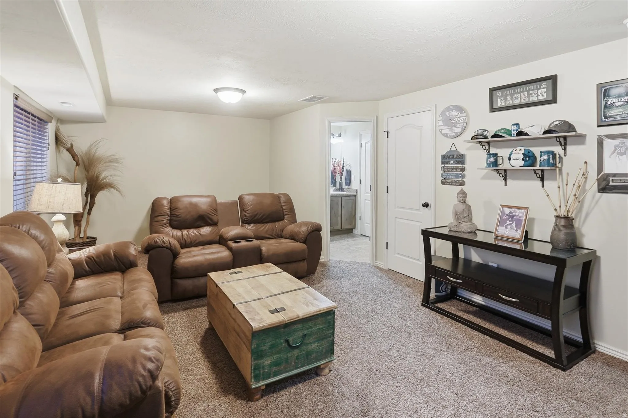 Carpeted recreation area with light-toned walls and ceiling, featuring a window with horizontal blinds and a ceiling-mounted light fixture