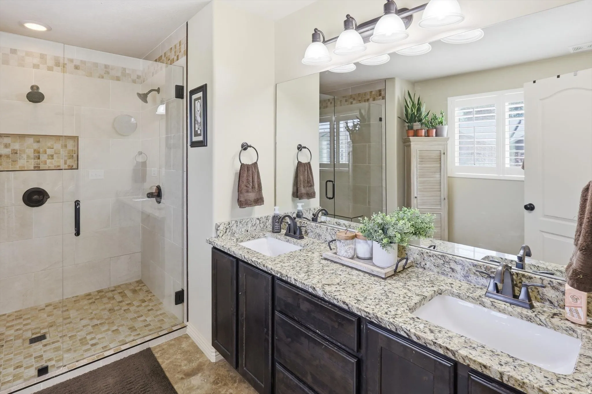 Bathroom featuring a glass-enclosed shower with tile surround and mosaic accent, dual vanity with granite countertop and undermount sinks, dark wood cabinetry, and a framed mirror with a four-light fixture