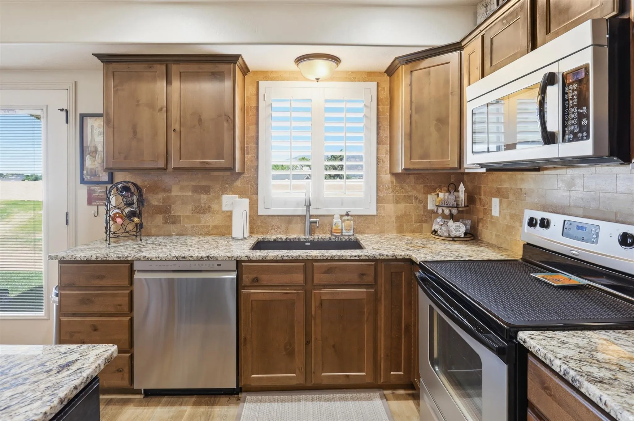 Kitchen featuring wood-finish cabinetry, speckled countertops, a mosaic tile backsplash, and stainless steel appliances including a dishwasher, sink, and oven with an overhead microwave