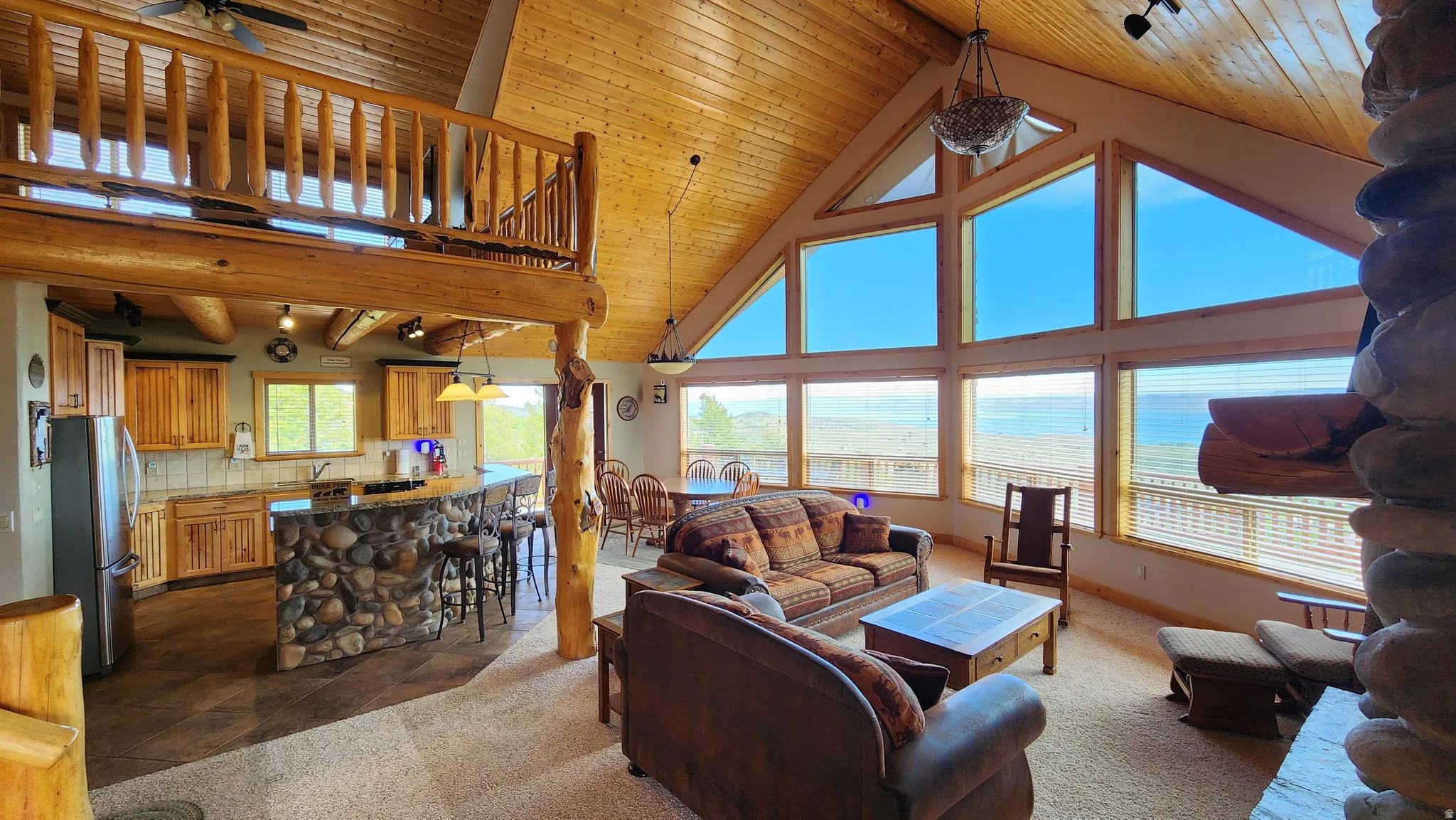 Open-concept living area featuring a vaulted wood-paneled ceiling, a stone-clad kitchen island with bar seating, and a loft with a wood railing