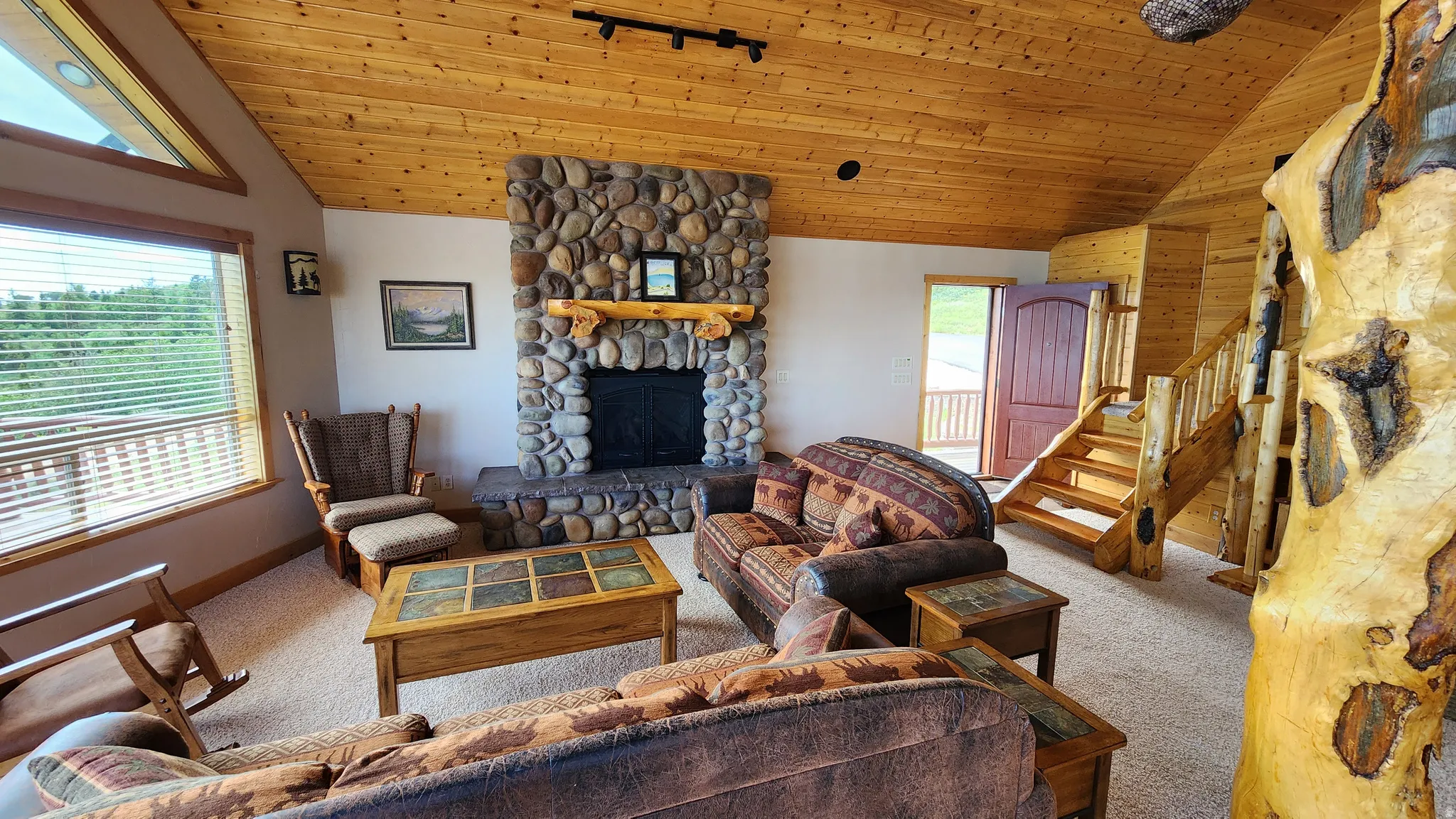 Living area featuring a floor-to-ceiling river rock fireplace, wood-plank ceilings, and a natural wood staircase