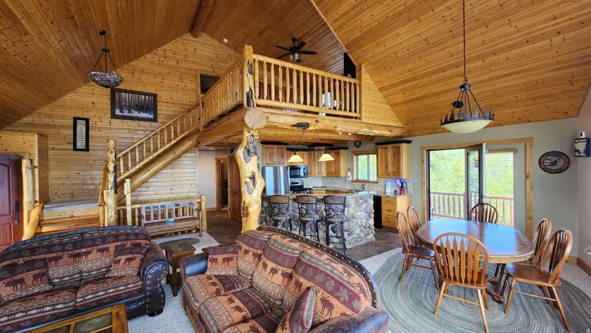 Open-concept living space featuring vaulted wood-paneled ceilings, a log staircase with a matching railing, a kitchen island with stone detailing, and a dining area with direct access to an outdoor balcony