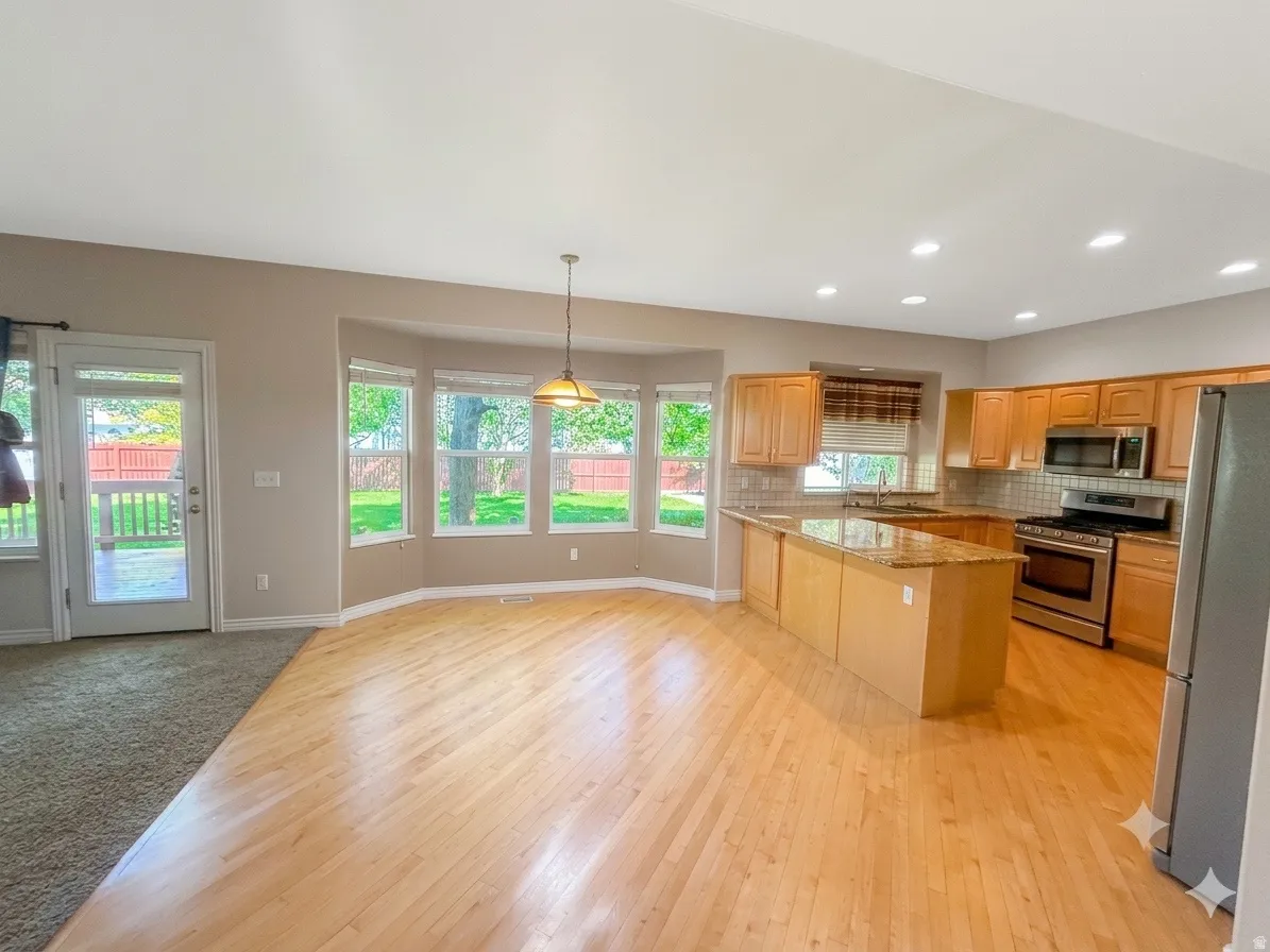 Open-concept kitchen and dining area featuring wood-finish flooring, a kitchen island with stone countertop, stainless steel appliances, and ample natural light from bay windows and a glass-paned door