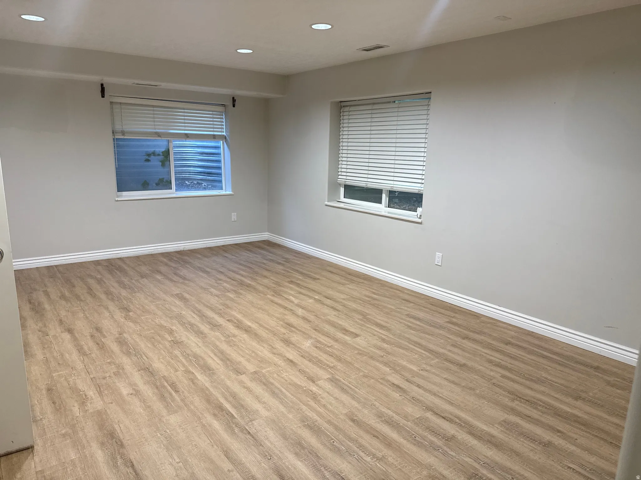 Spacious room featuring wood-finish flooring, light grey painted walls, recessed lighting, and two windows with horizontal blinds