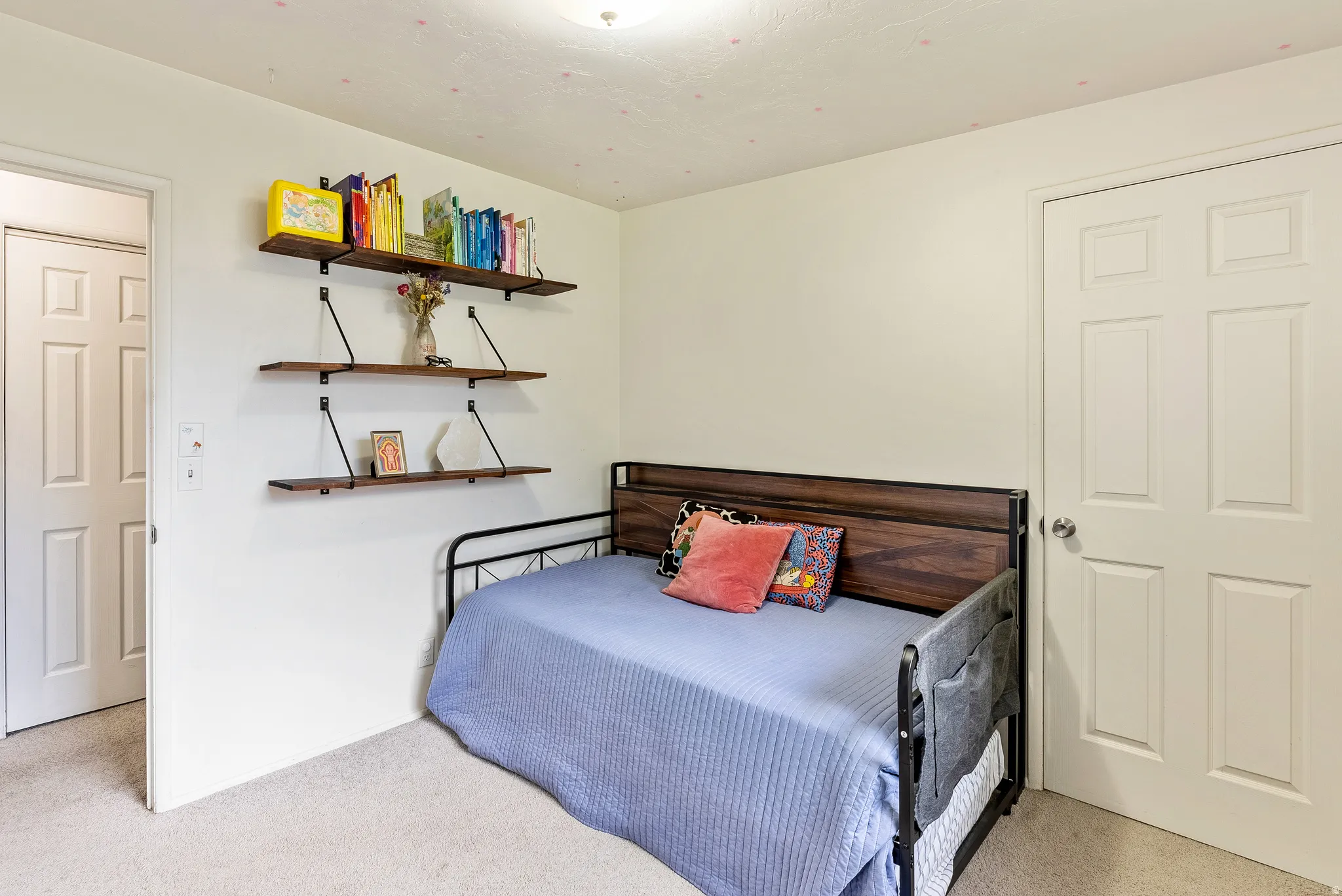 Carpeted room with a ceiling light fixture, three wood-finish wall-mounted shelves with black metal brackets, and two white paneled doors