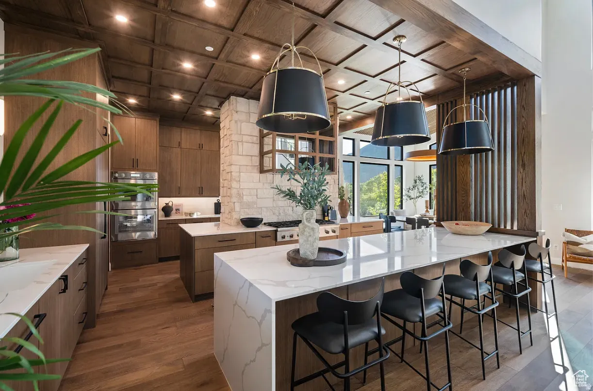 Gourmet kitchen featuring a large waterfall island with seating, stone veneer accent wall, wood-finish coffered ceiling, stainless steel double ovens, and expansive windows