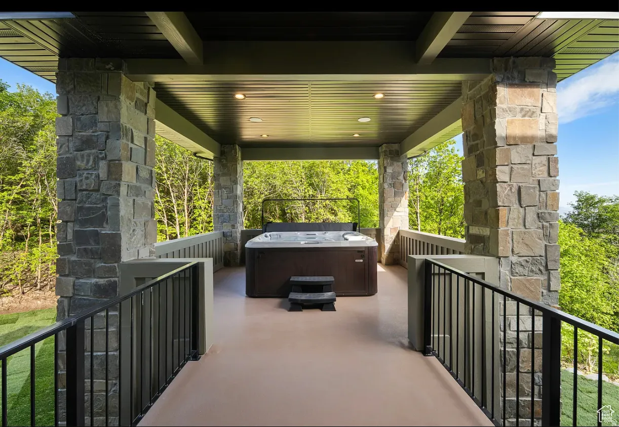 Covered outdoor patio featuring a built-in hot tub, recessed lighting, stone-clad columns, and black metal railings
