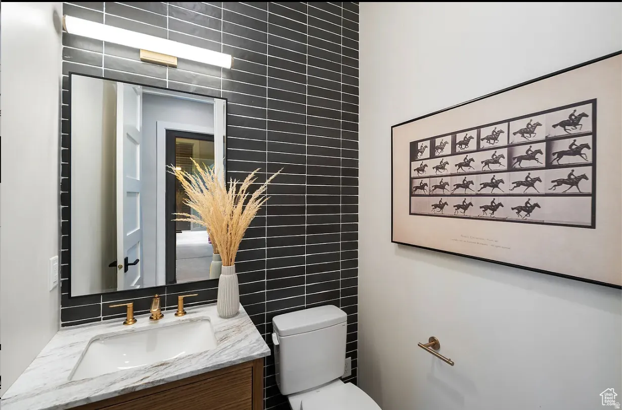 Powder room featuring a dark tiled accent wall, a rectangular mirror with a modern vanity light, a wood-finish vanity with a white countertop and undermount sink, and brass-tone fixtures