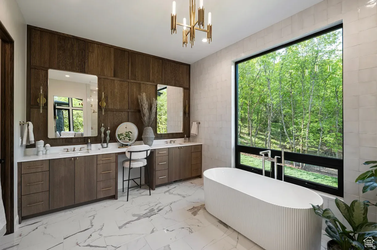 Bathroom featuring a freestanding soaking tub, a large picture window, a double vanity with stone countertops, wood-finish cabinetry, and marble-look tile flooring