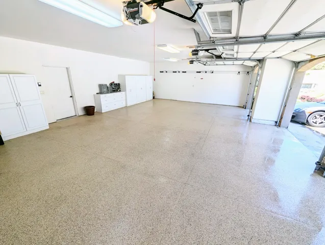 Spacious garage featuring epoxy flooring, overhead lighting, multiple built-in storage cabinets, and a white access door