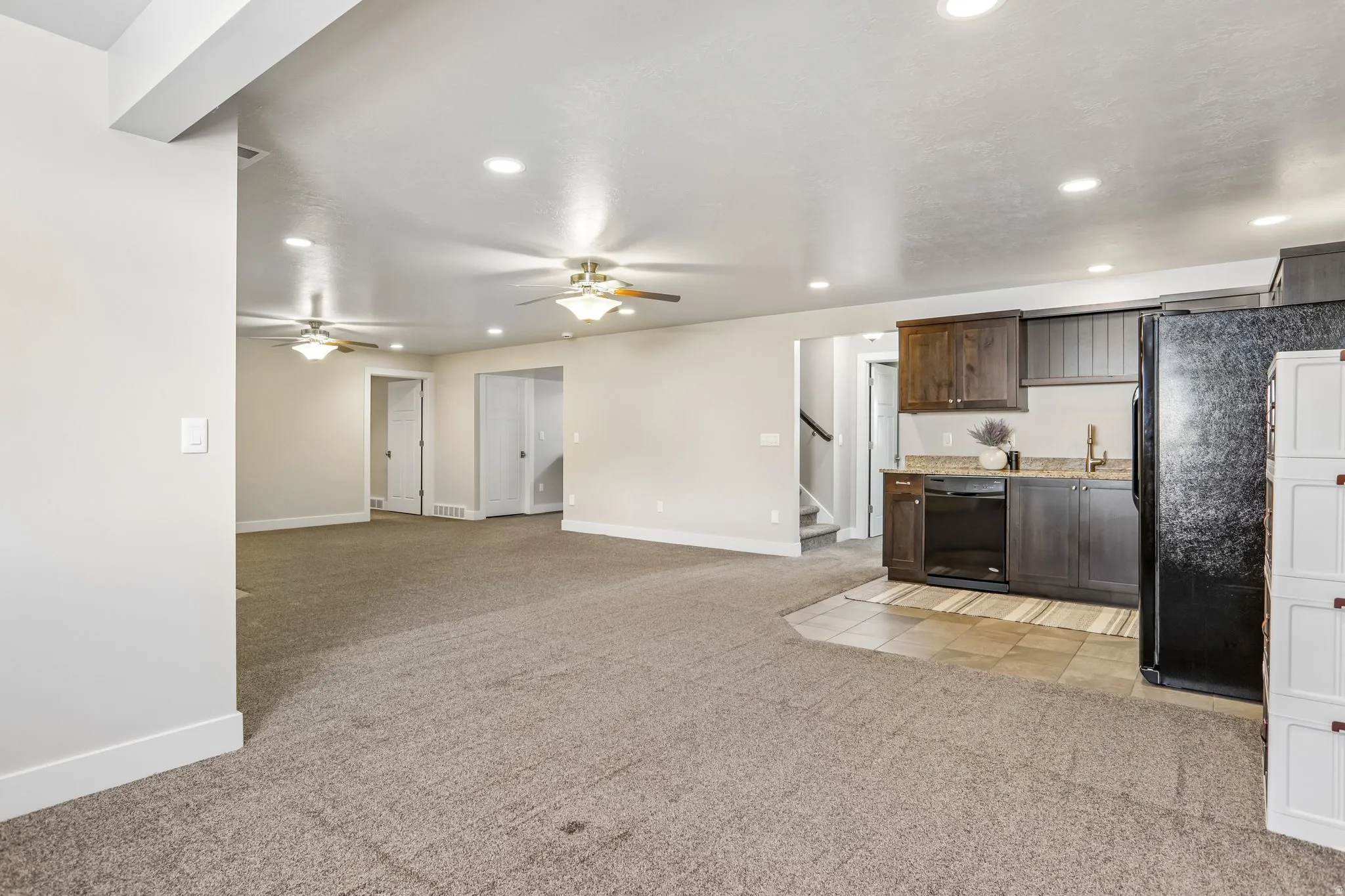 Spacious open-concept room featuring a functional kitchenette with dark wood-finish cabinetry, a black refrigerator, and a built-in black dishwasher
