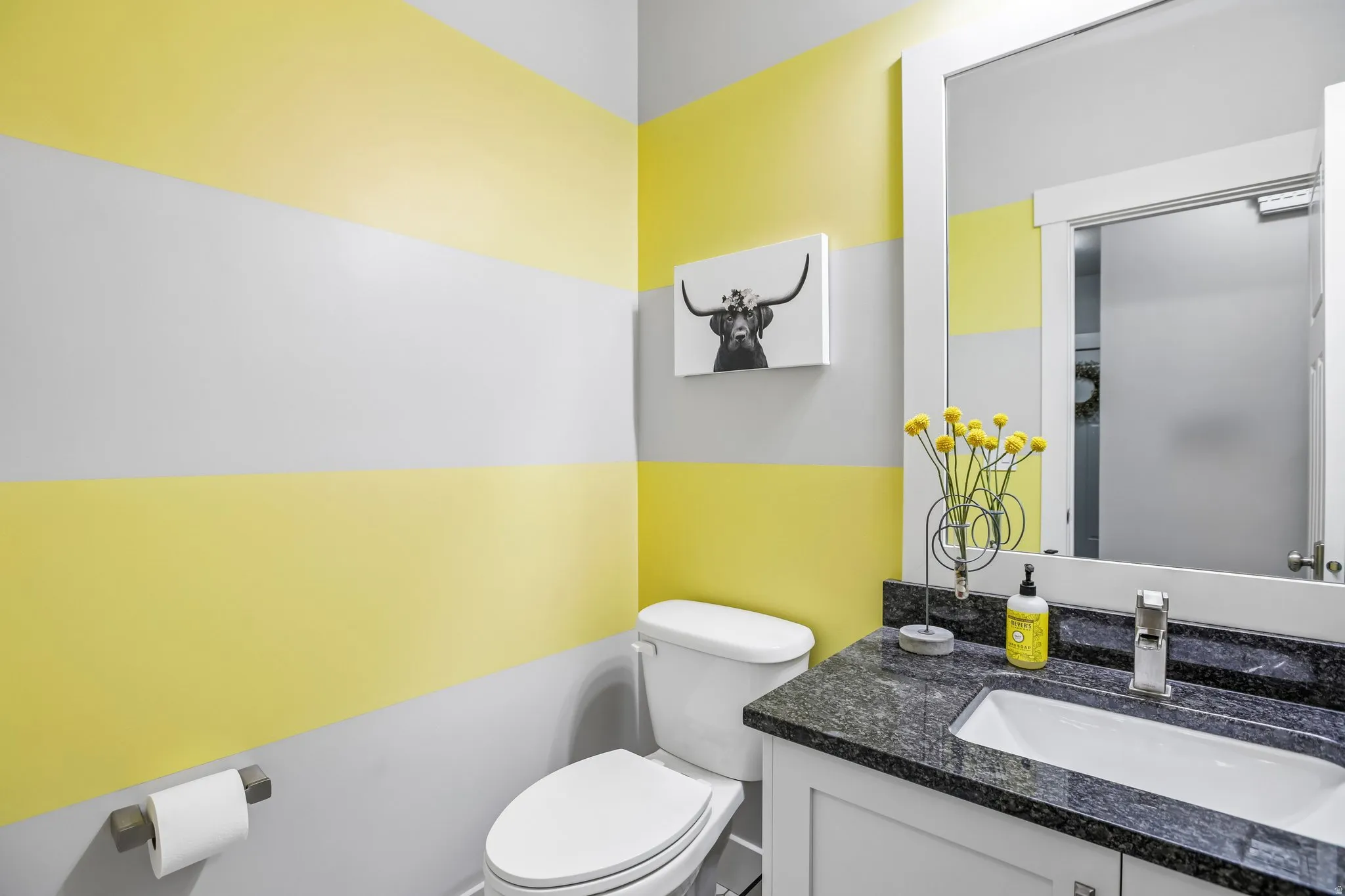 Bathroom featuring horizontal striped walls, a white vanity with a dark granite countertop, an undermount sink, and a framed wall mirror