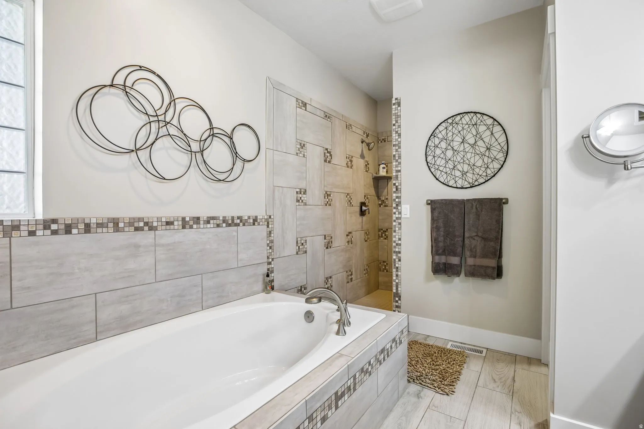 En suite bathroom featuring a built-in soaking tub with custom tile surround, a tile-enclosed shower with two heads, a frosted glass window, and heated plank flooring flooring