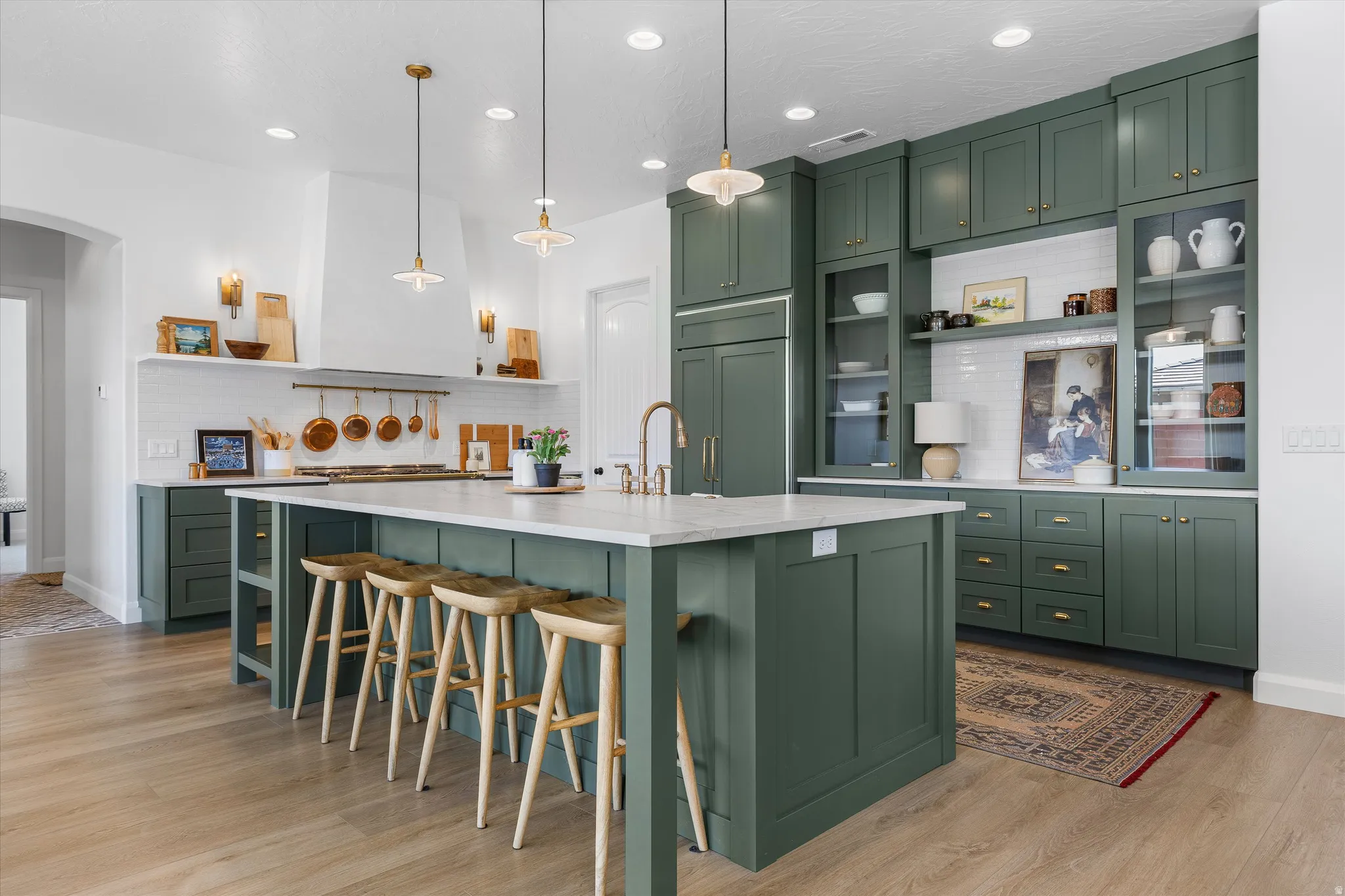 Spacious kitchen featuring an expansive central island with seating, ample green cabinetry with gold hardware, integrated range hood, white subway tile backsplash, and light wood-finish flooring