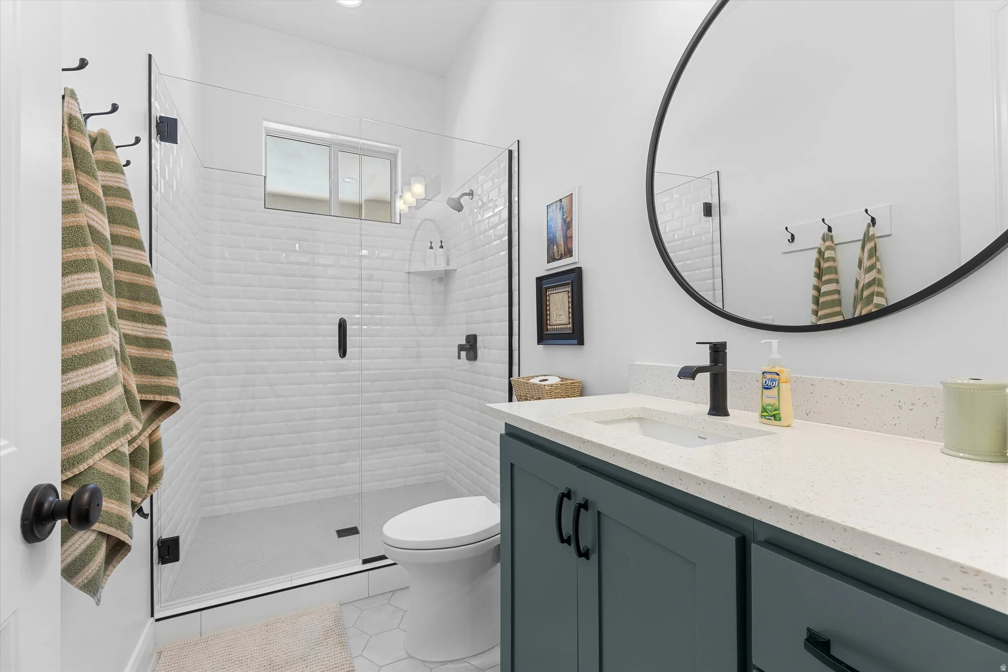 Modern bathroom featuring a glass-enclosed shower with subway tile surround, a dark teal vanity with a white countertop, a black-framed circular mirror, and black hardware accents throughout