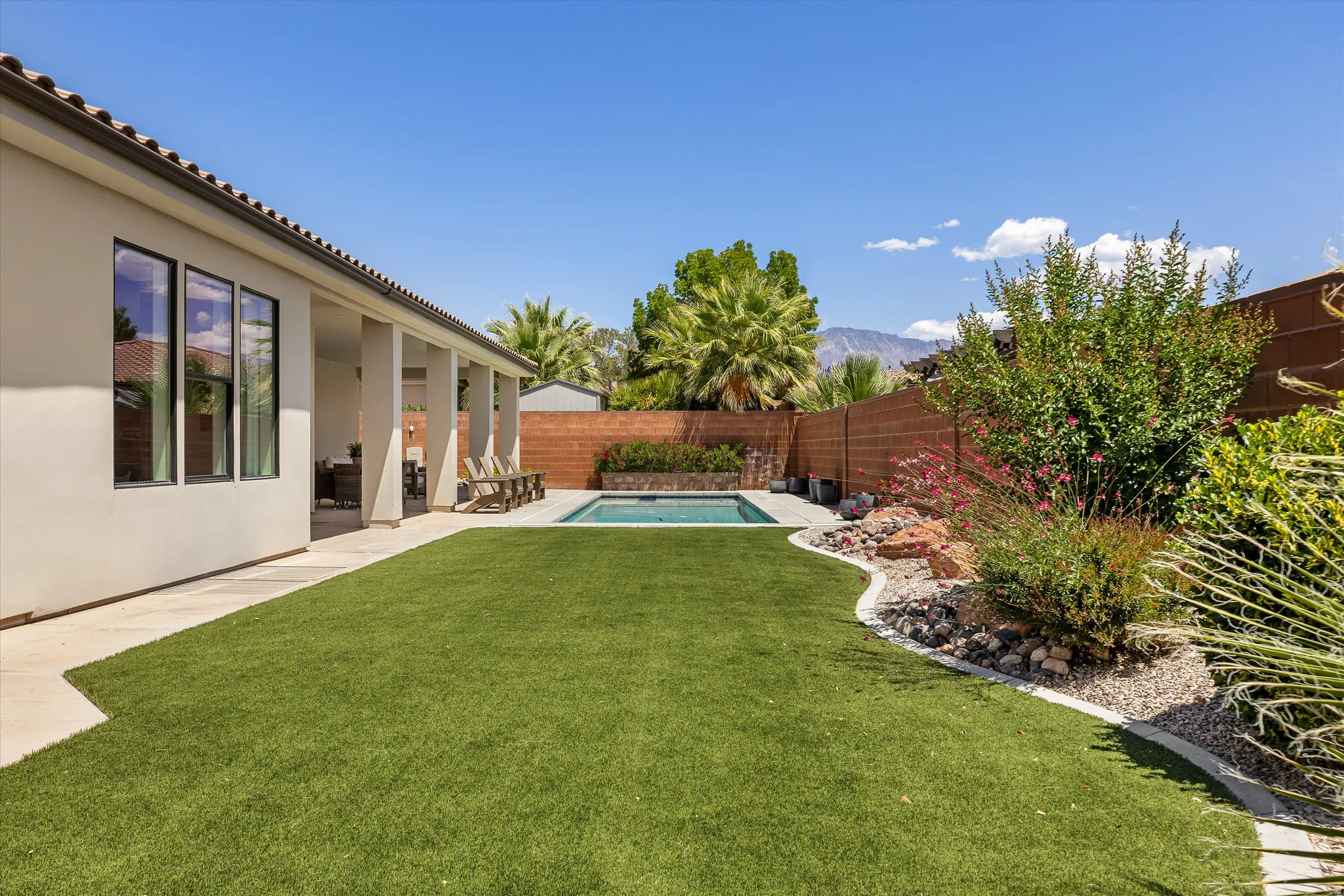 Expansive backyard featuring a rectangular swimming pool, covered patio, artificial turf, and desert landscaping with mature trees