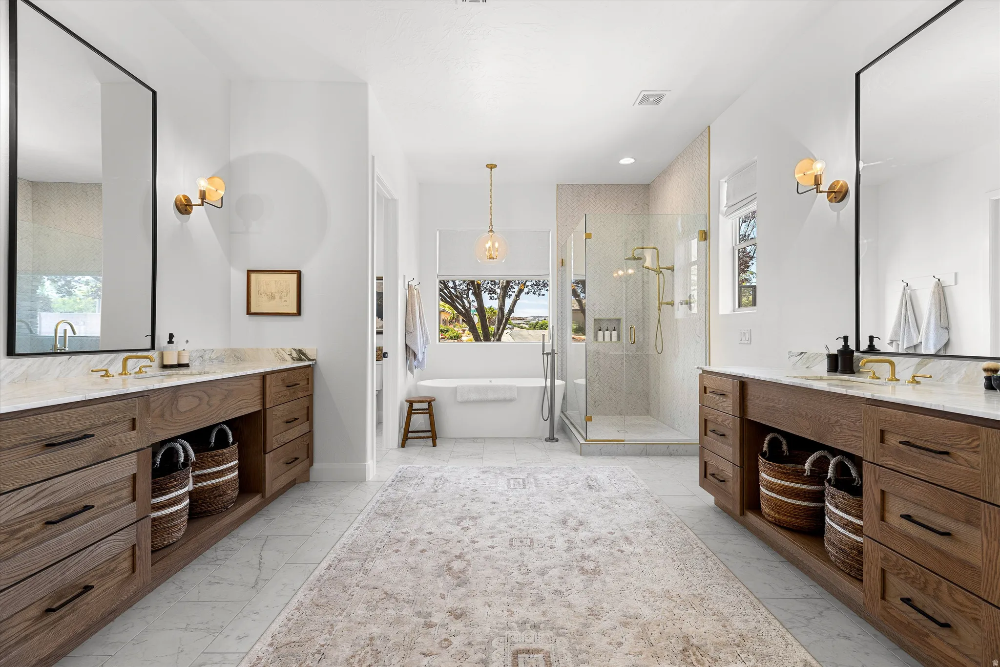 Spacious bathroom featuring dual wood-finish vanities with stone countertops, a freestanding tub, and a glass-enclosed shower with gold-tone fixtures