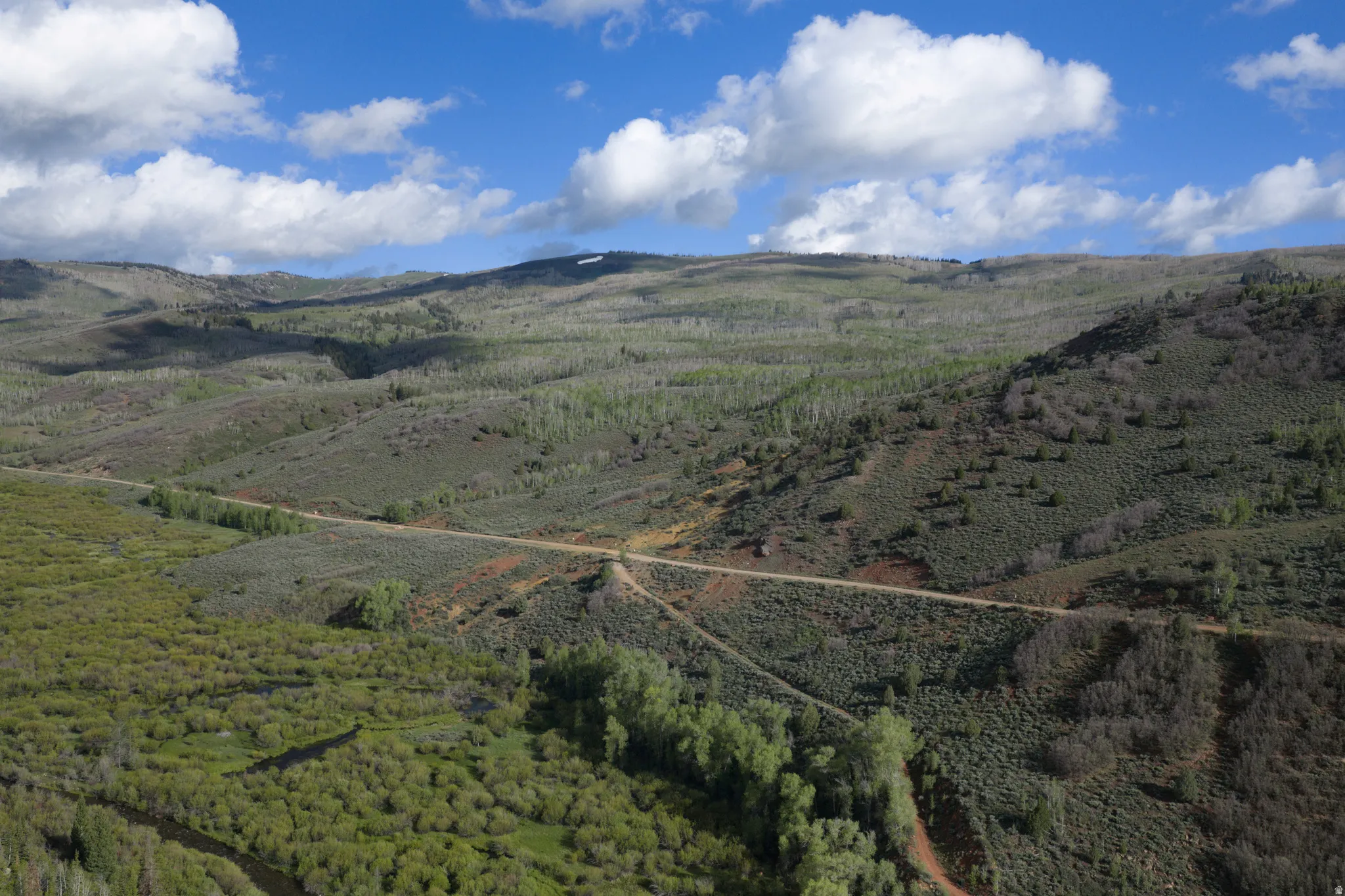 Expansive mountainous terrain featuring diverse vegetation, a winding dirt road, and a visible stream