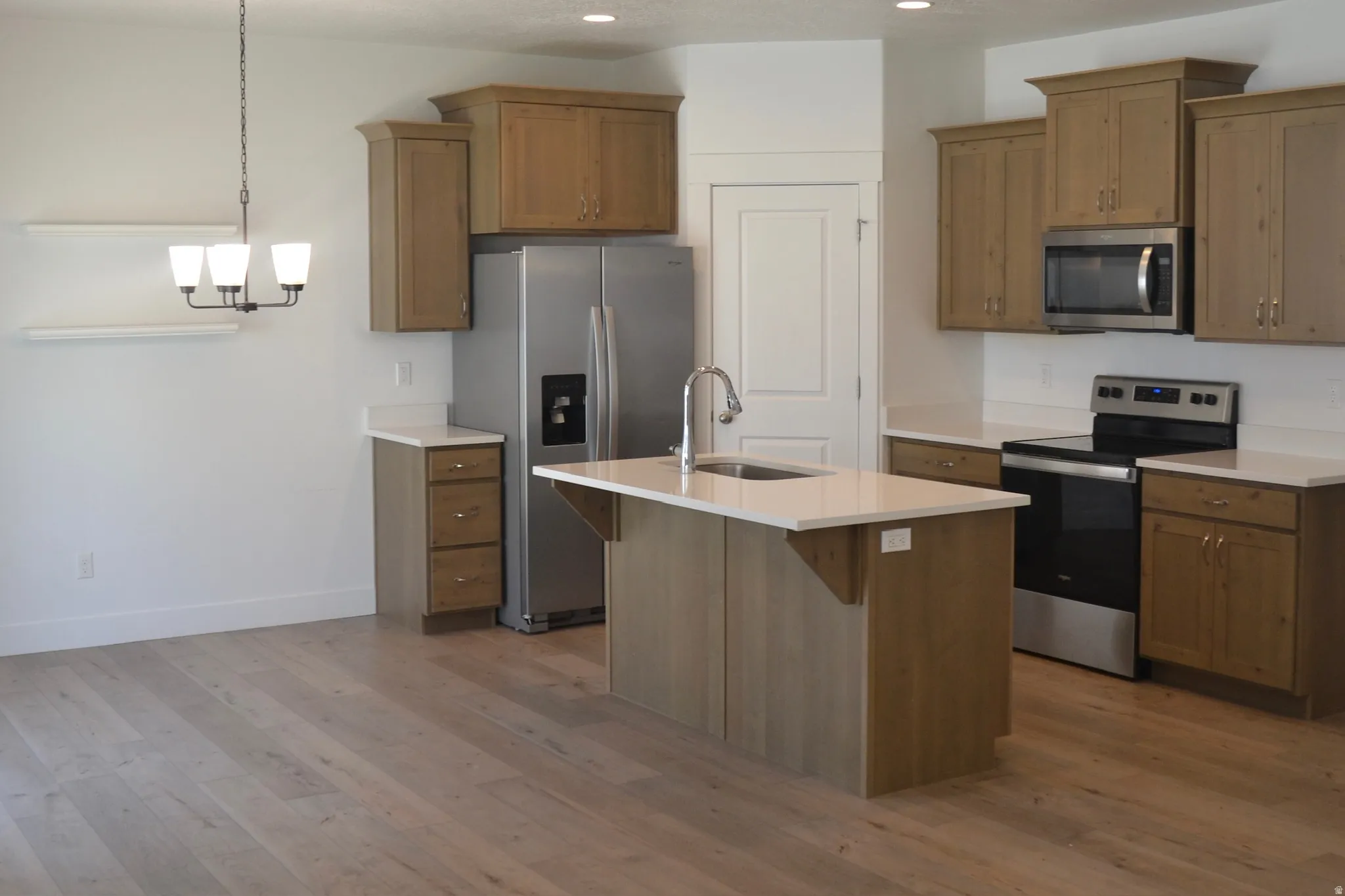 Kitchen space featuring wood-finish flooring, light cabinetry, stainless steel appliances, a center island with sink, and light-toned countertops
