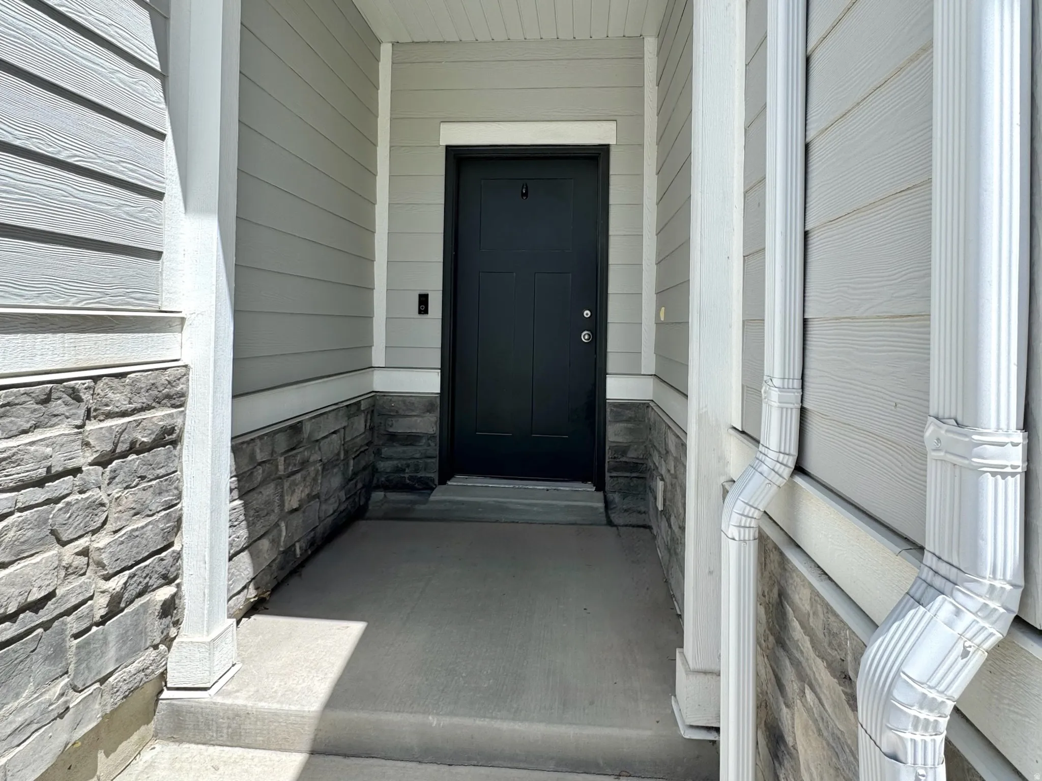 Inviting front entryway featuring a solid black door, concrete porch, stone veneer siding, horizontal panel siding, and white trim accents