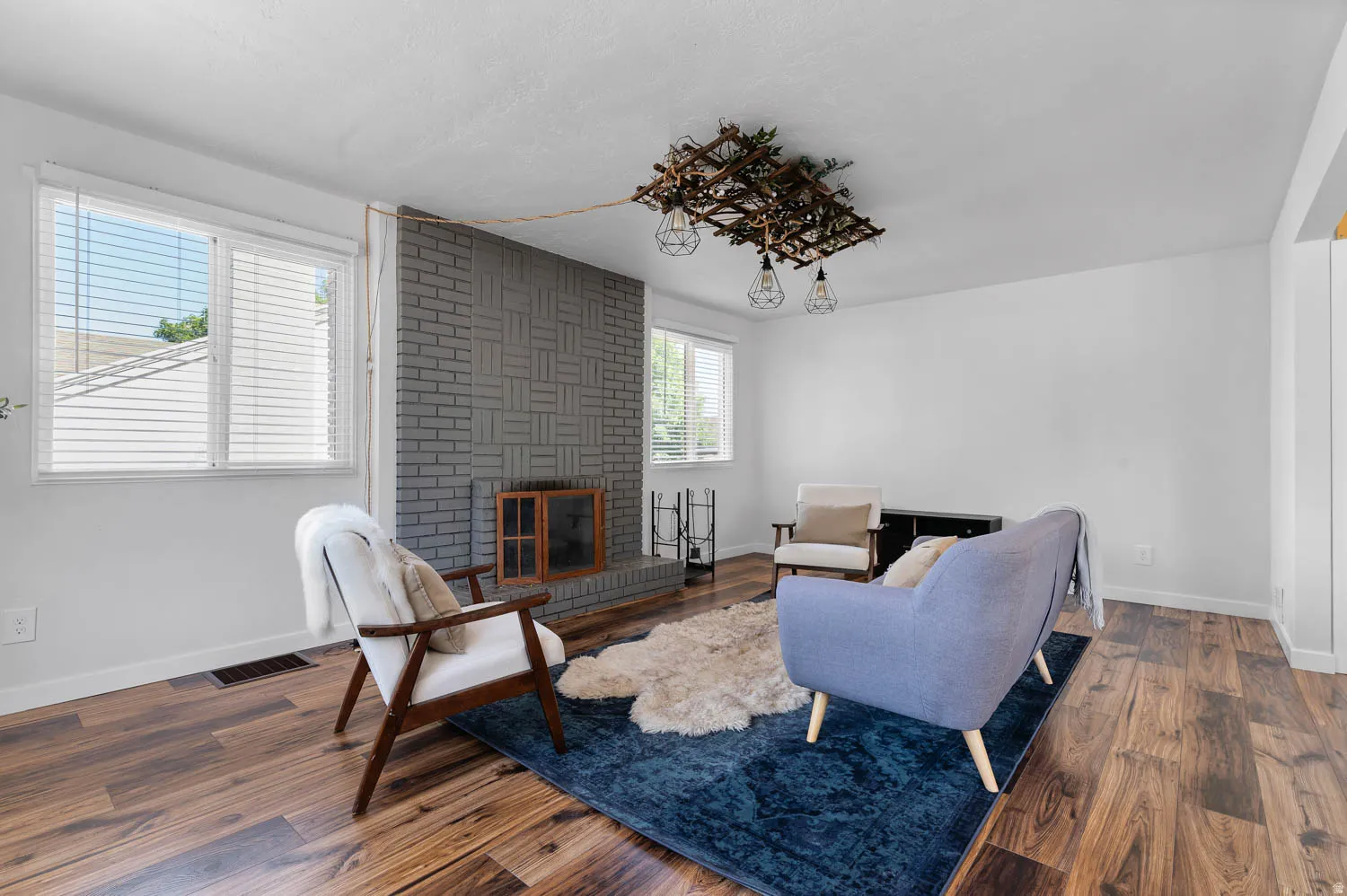 Living area featuring wood-finish flooring, a painted brick fireplace, and dual windows with blinds
