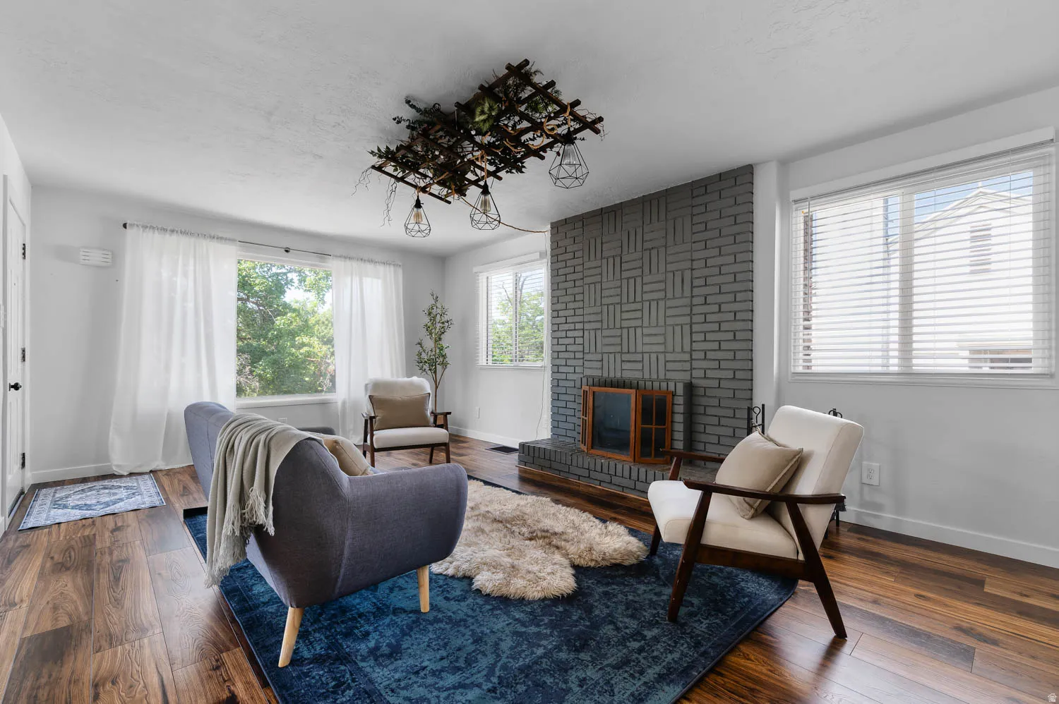 Living area featuring wood-finish flooring, a painted brick fireplace, a decorative ceiling light fixture, and multiple windows with natural light