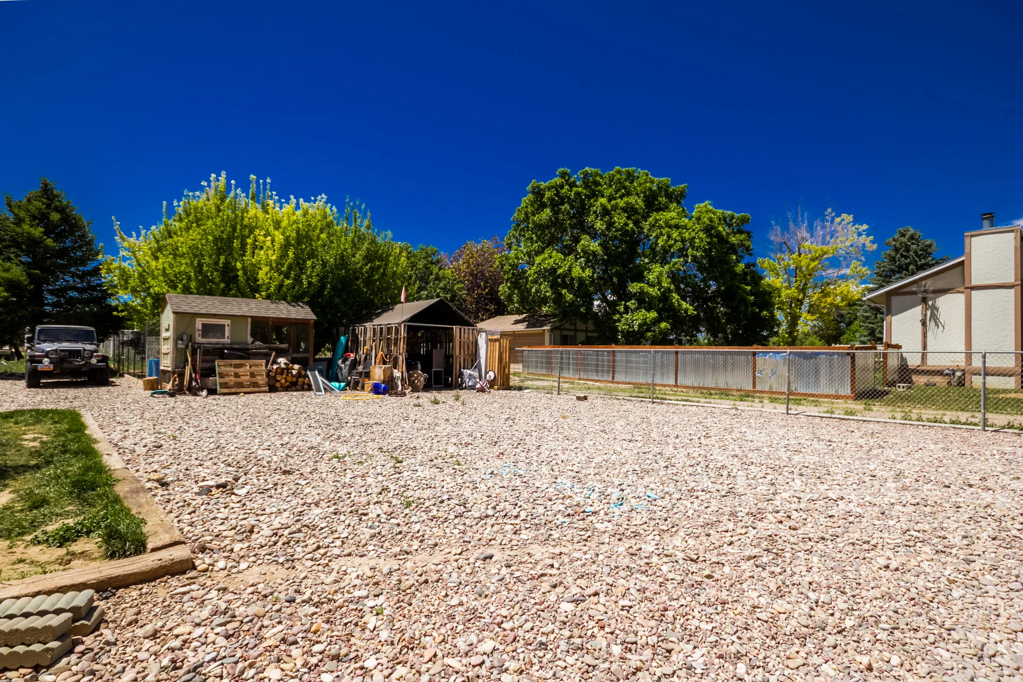 Expansive gravel yard featuring multiple outbuildings, mature trees, and a chain-link fence