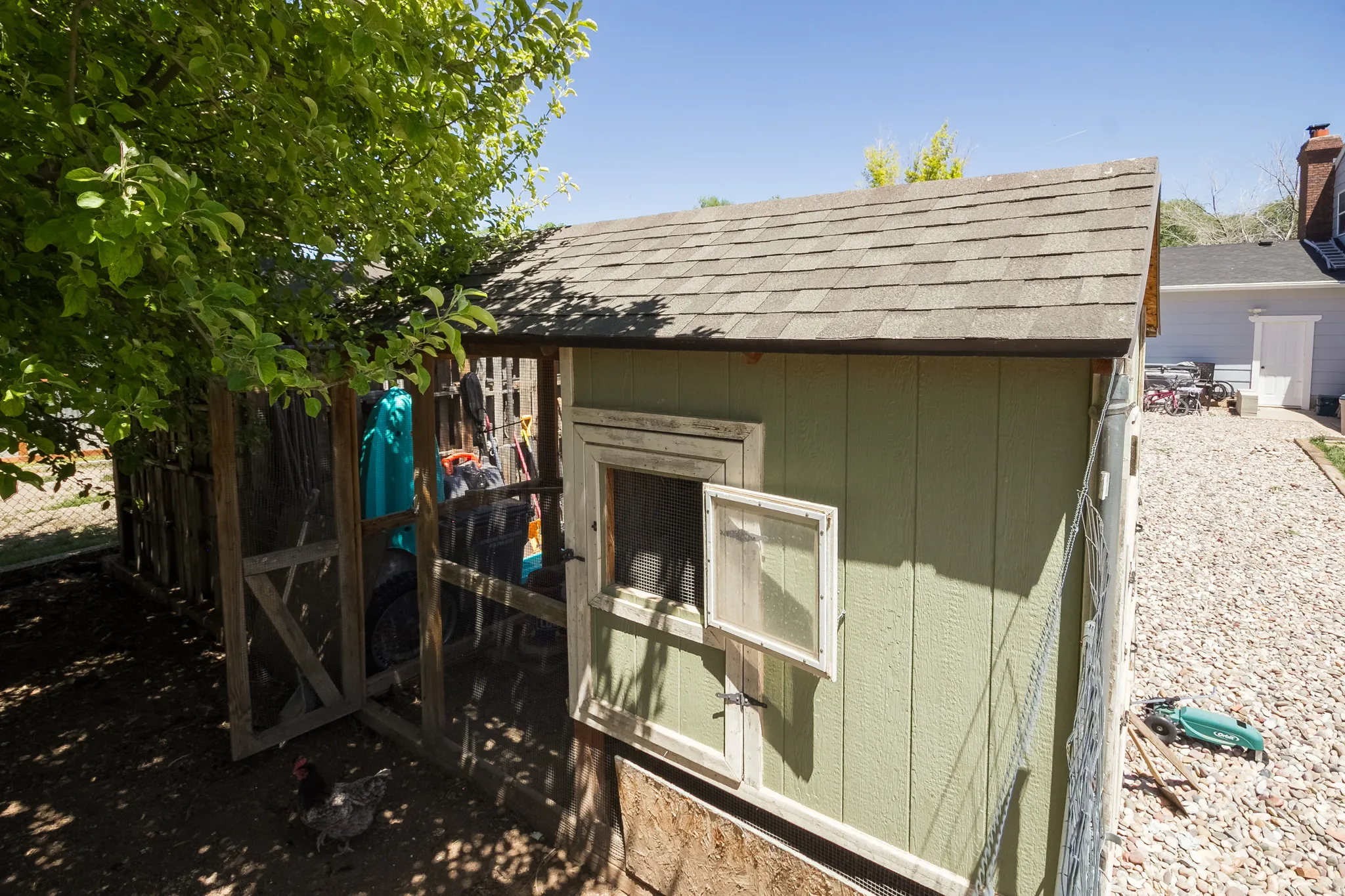 Green exterior utility structure with a shingled roof, featuring a screened window and an attached fenced enclosure with a wooden frame