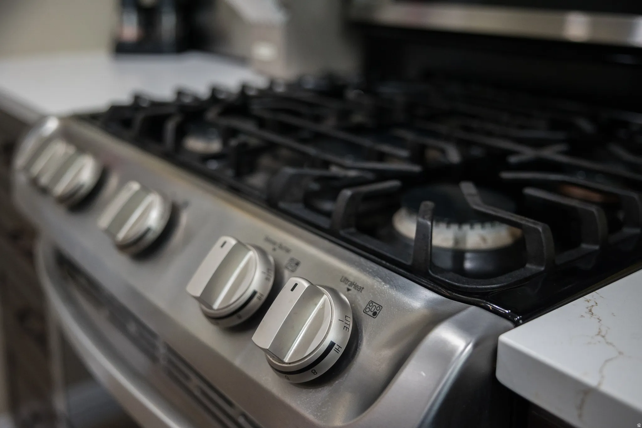 Stainless steel gas range featuring multiple burners and grates, brushed metal control knobs, and integration with a light-toned countertop with veining
