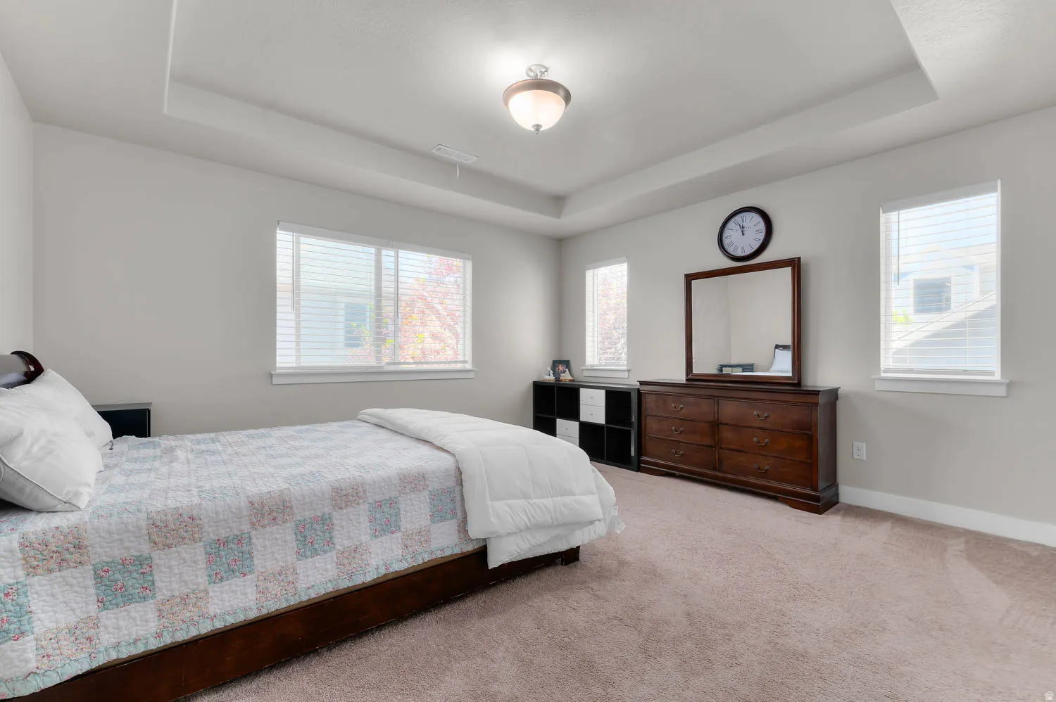 Carpeted bedroom featuring a tray ceiling with a semi-flush mount light fixture, two windows with blinds, and light-colored walls