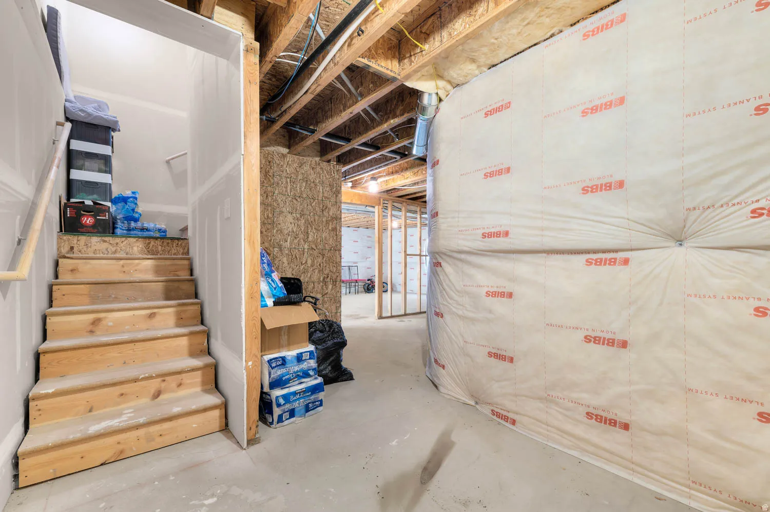Unfinished basement space featuring exposed wooden ceiling joists, concrete flooring, and a wooden staircase with a handrail