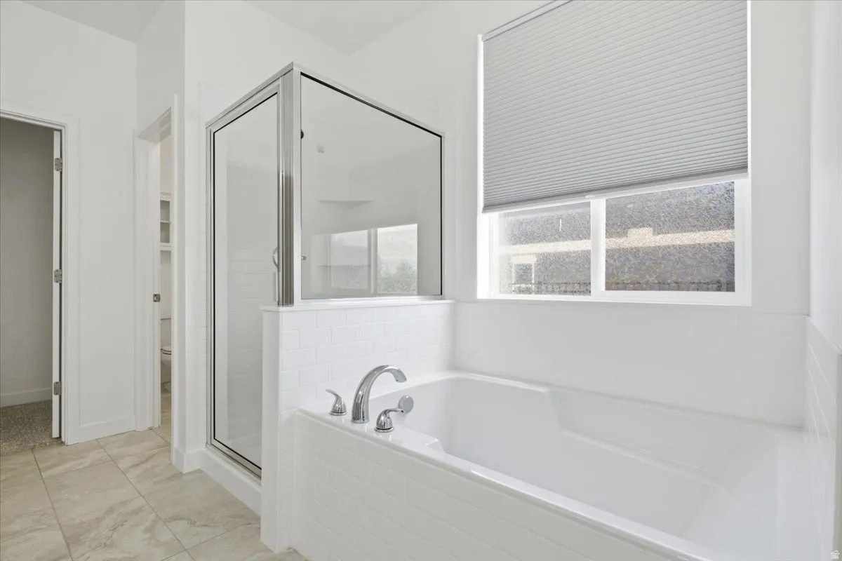 Spacious bathroom featuring a built-in soaking tub with chrome fixtures, a separate glass-enclosed shower, a large window with privacy blinds, and light-toned tile flooring