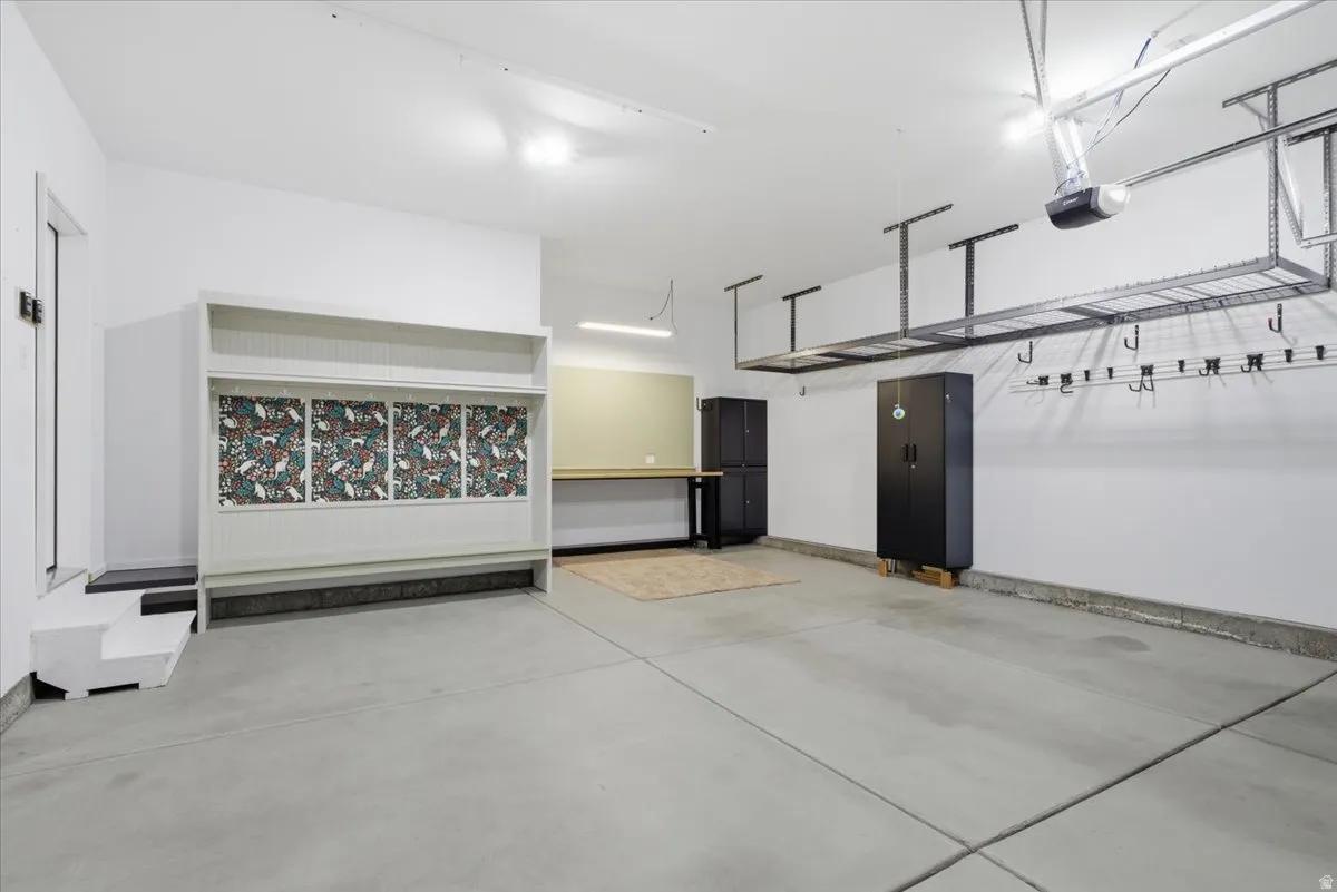 Spacious garage featuring a concrete floor, built-in storage unit with patterned backing, wall-mounted workbench, overhead storage racks, and white walls