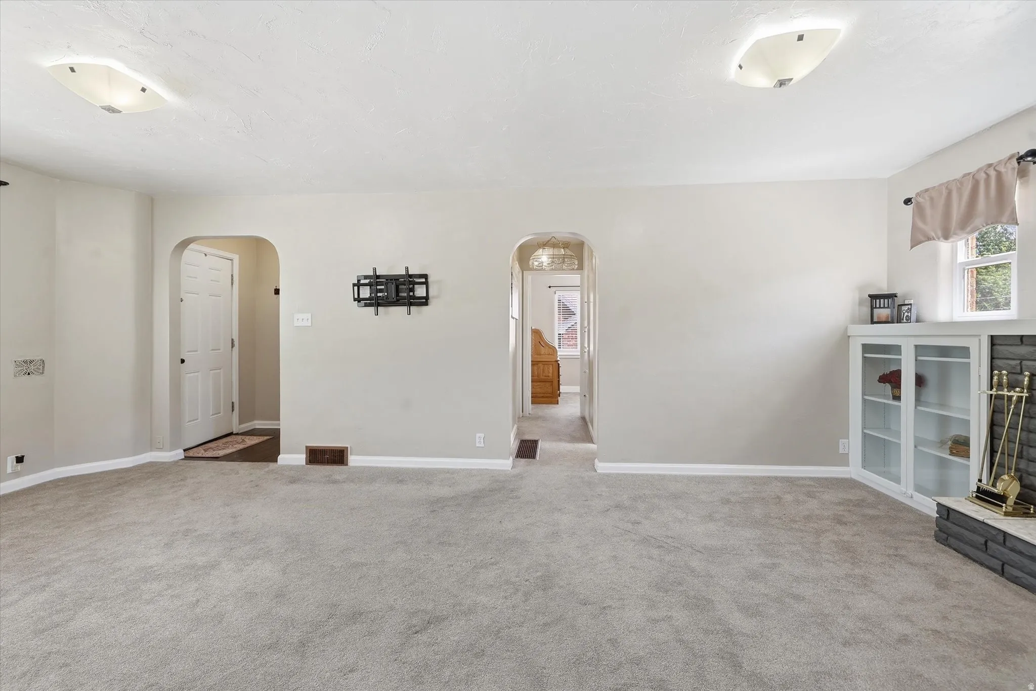 Spacious carpeted room featuring arched doorways, ceiling-mounted light fixtures, a corner built-in cabinet with glass doors, and a brick-faced fireplace hearth