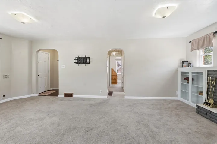 Spacious carpeted room featuring arched doorways, ceiling-mounted light fixtures, a corner built-in cabinet with glass doors, and a brick-faced fireplace hearth