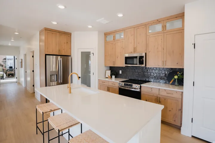 Contemporary kitchen featuring a large central island with a white countertop and integrated sink, light-toned wood cabinetry, stainless steel appliances, and a dark tile backsplash
