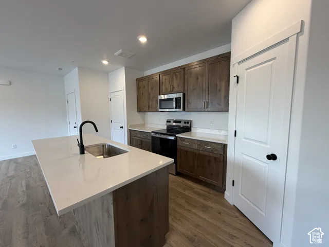 Kitchen featuring an island with an integrated sink, wood-finish flooring, stainless steel appliances, wood cabinetry, and recessed lighting