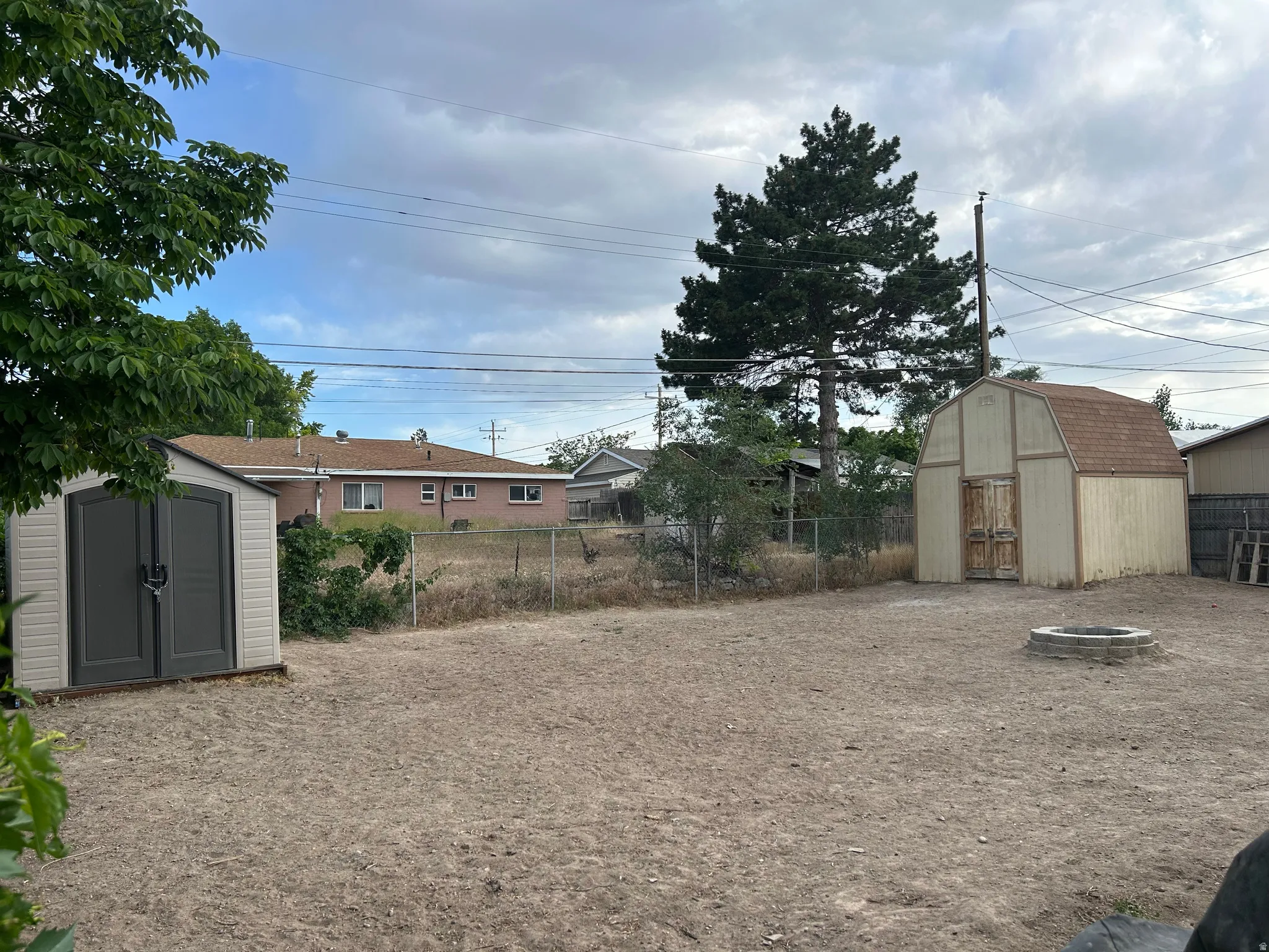 Expansive backyard featuring two storage sheds, a chain-link fence, and a built-in fire pit