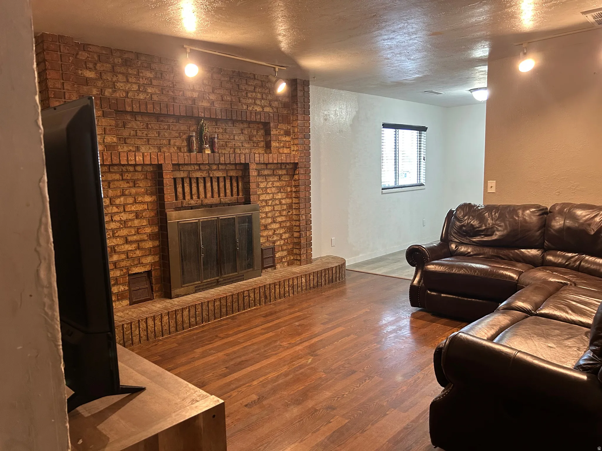 Spacious living area featuring wood-finish flooring, a prominent brick fireplace with hearth, and track lighting