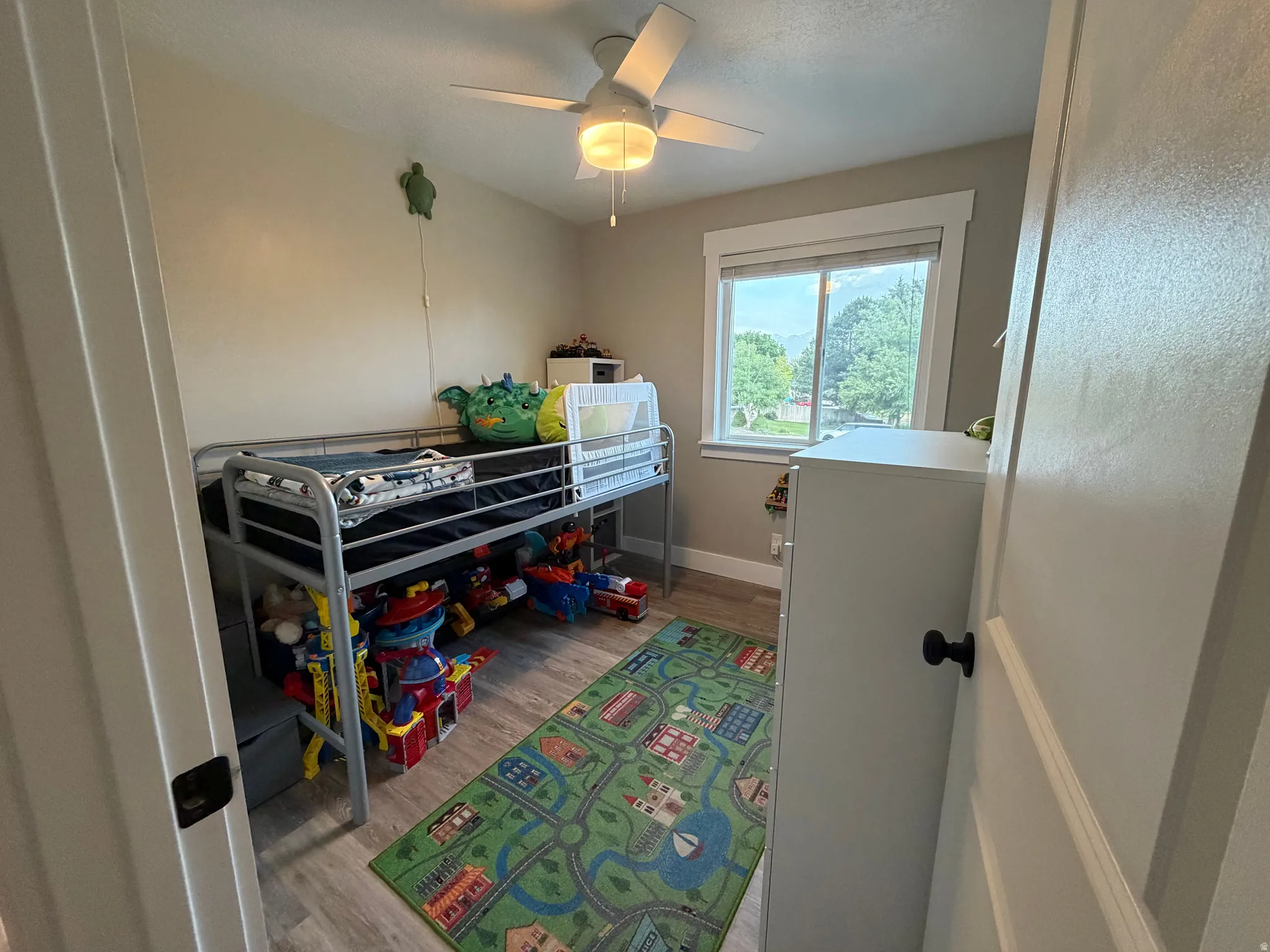 Room featuring a ceiling fan with light fixture, window with exterior landscape, wood-finish flooring, and white trim