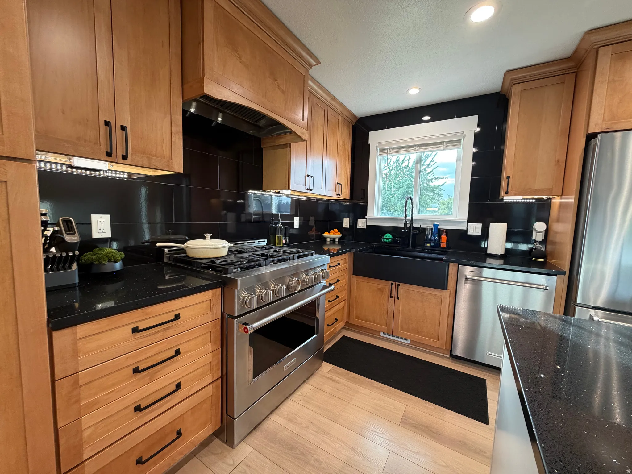 Modern kitchen featuring light wood cabinetry with black hardware, dark granite countertops, a black tile backsplash, stainless steel appliances, and wood-finish flooring