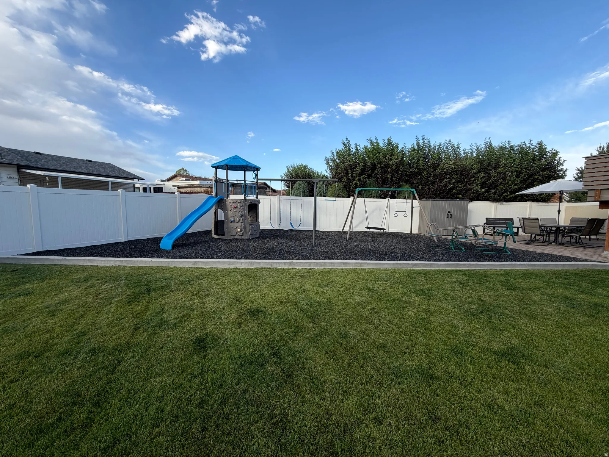Expansive backyard featuring a manicured lawn, white vinyl fencing, a dedicated play area with rubber mulch surfacing, and a stone-look patio with paver flooring