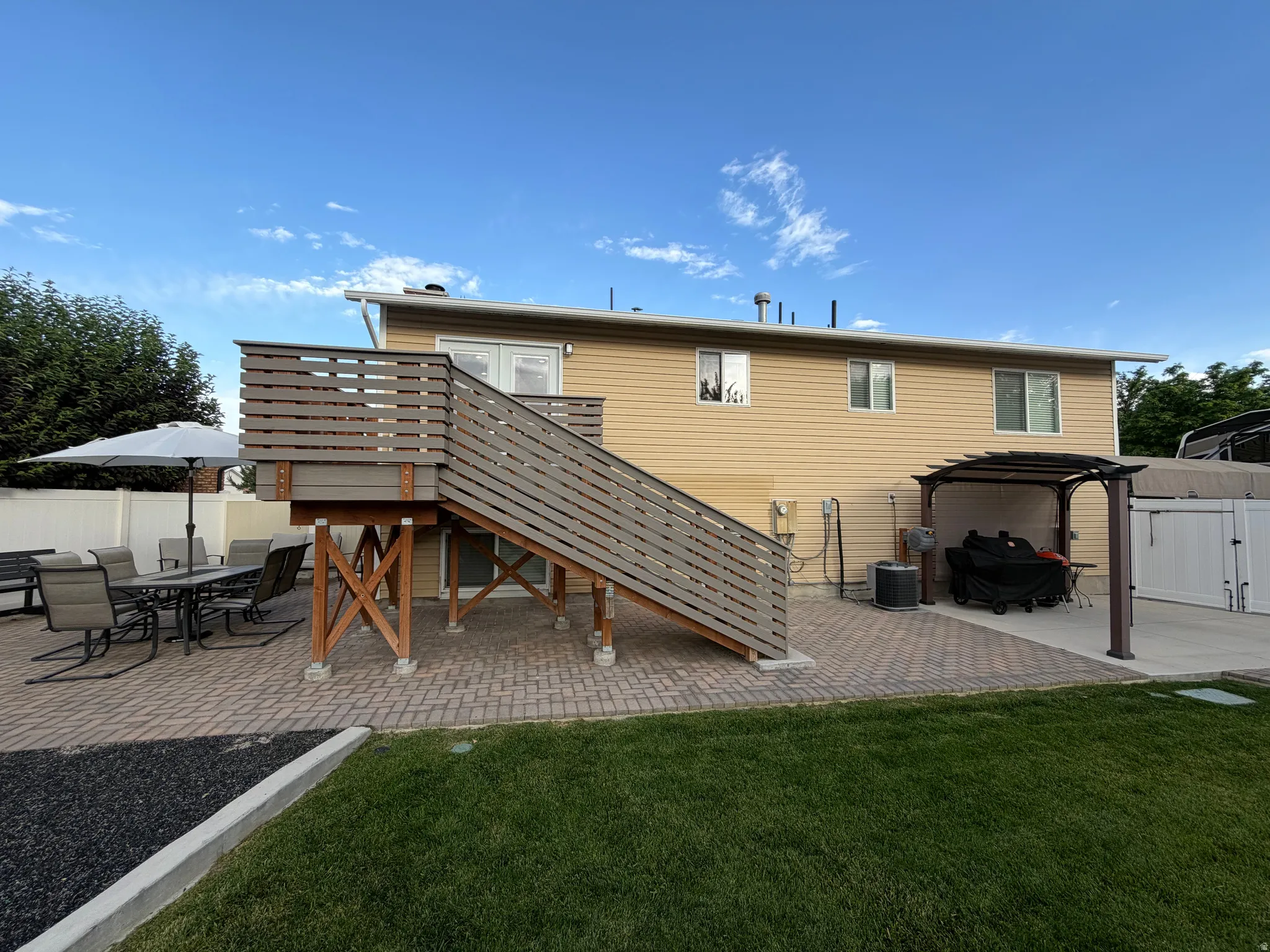 Multi-level outdoor living space featuring a raised deck with horizontal slat railings, a herringbone pattern paver patio, a separate concrete patio area, a pergola, and a vibrant green lawn