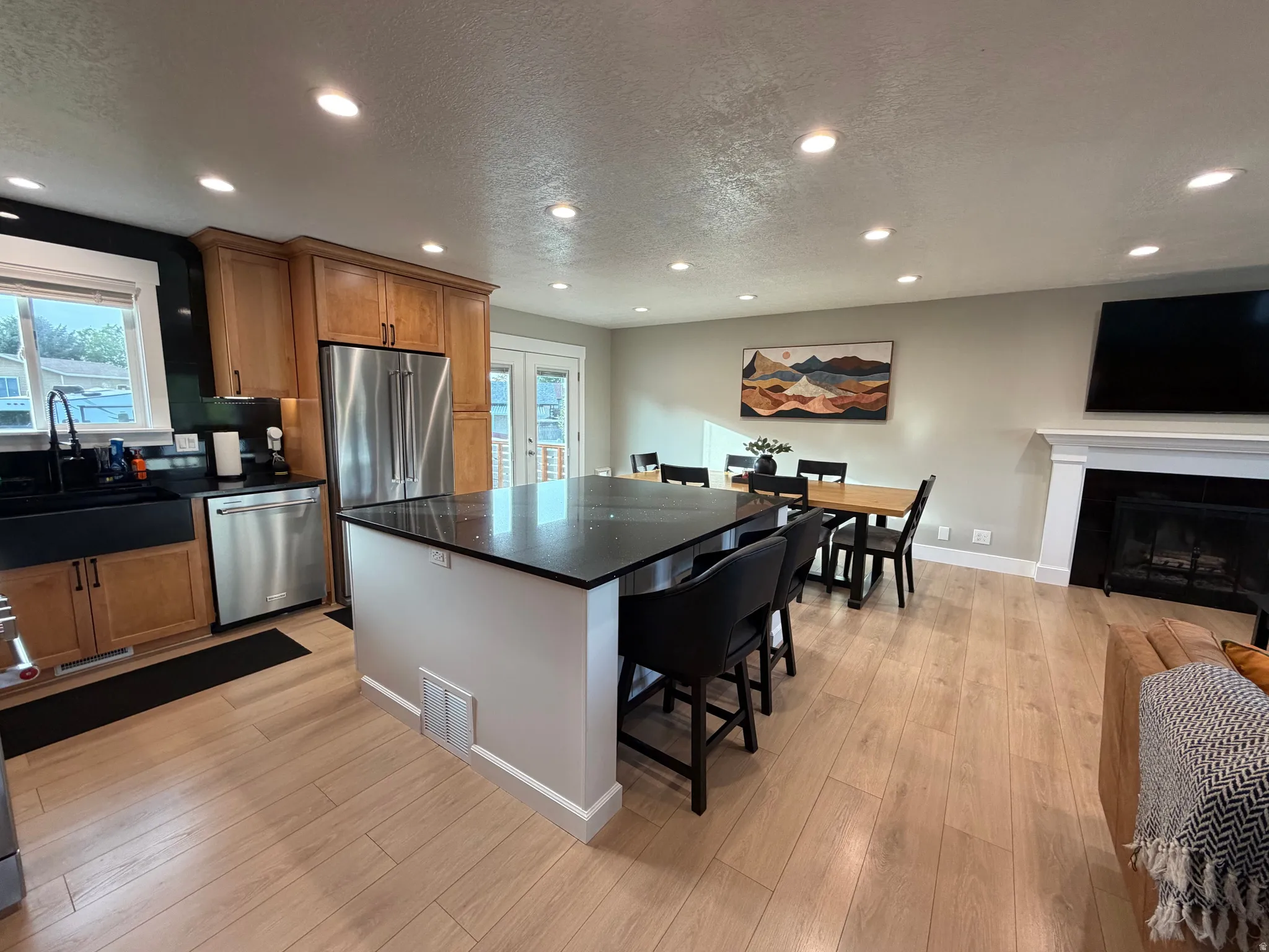 Open-concept living area featuring wood-finish flooring, a kitchen island with dark countertops, stainless steel appliances, and a fireplace with a white mantel