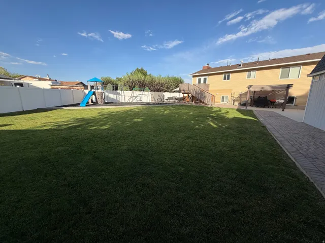 Expansive backyard featuring a manicured lawn, a full white vinyl privacy fence, a paver pathway, a concrete patio, and a covered pergola