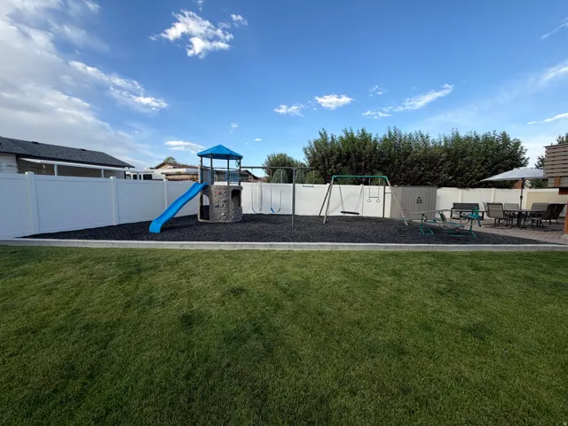 Expansive backyard featuring a manicured lawn, white vinyl fencing, a dedicated play area with rubber mulch surfacing, and a stone-look patio with paver flooring