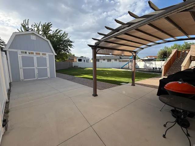 Expansive concrete patio area featuring a pergola with a retractable shade canopy and a separate detached shed with double doors